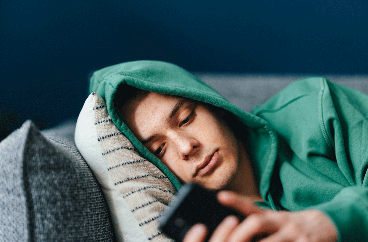 Ein deprimierter Jugendlicher im Kapuzenpullover liegt auf dem Sofa und schaut auf das Handy
