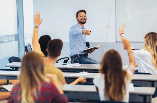 Präsenzunterricht mit Erwachsenen: Der Lehrer zeigt auf zwei Studierende, die ihre Hände hochheben und eine Frage zu stellen.