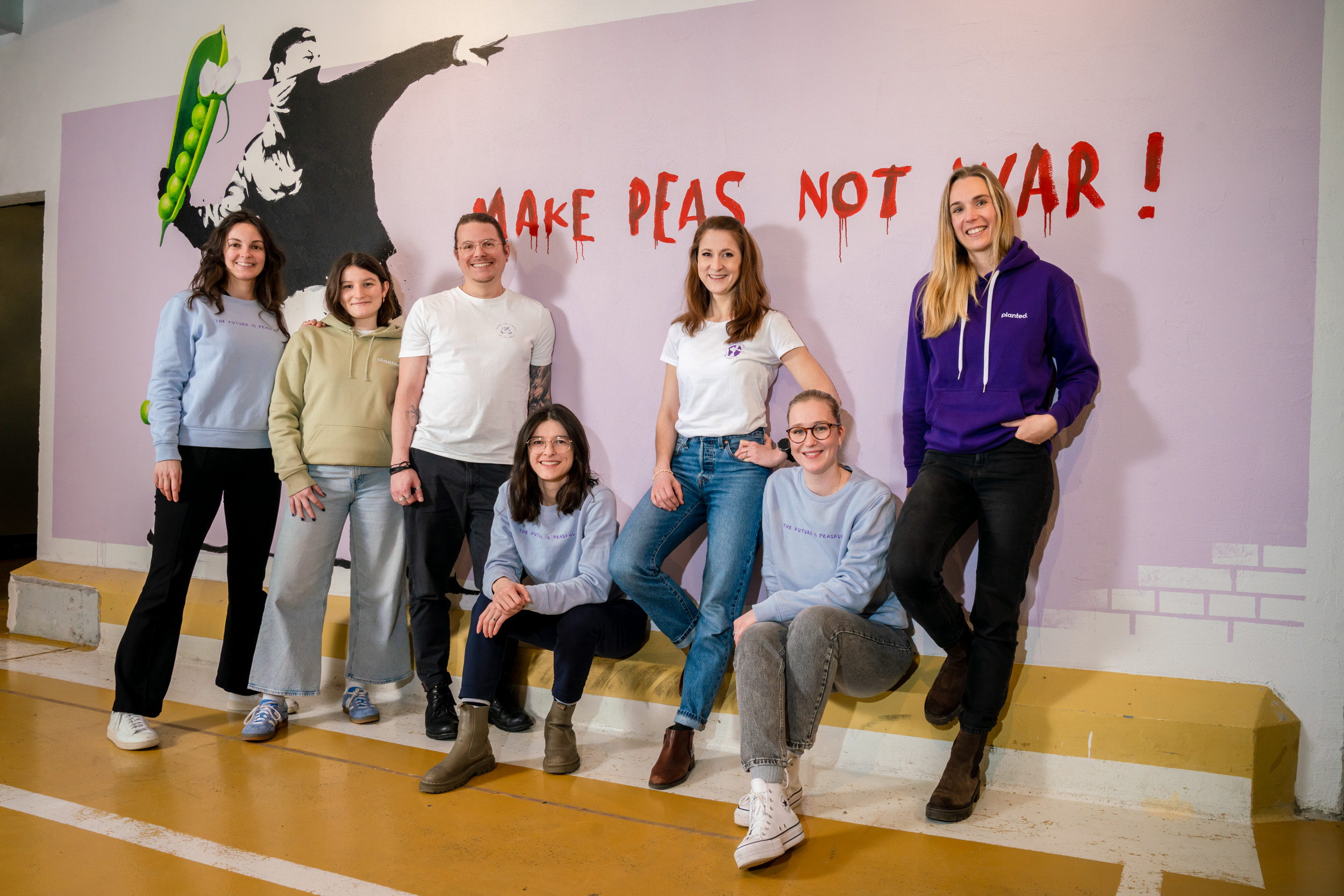 Das Bild zeigt das HR-Team von Planted, eine Gruppe von jungen Menschen in lockerer, modischen Kleidung vor einem Banksy-Kunstwerk mit der Graffiti-Aufschrift "Make peas not war!"