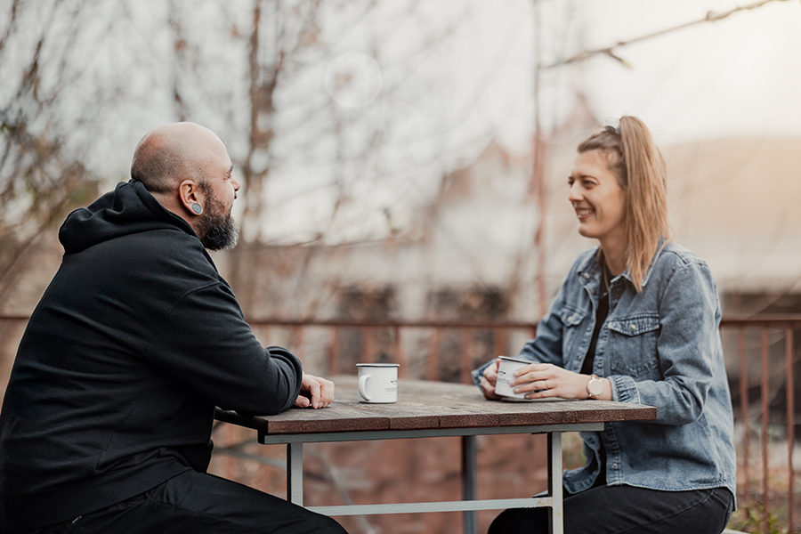 Ein Mann und eine Frau in lockerer Kleidung sitzen beim Kaffee an einem Tisch auf einer Terrasse