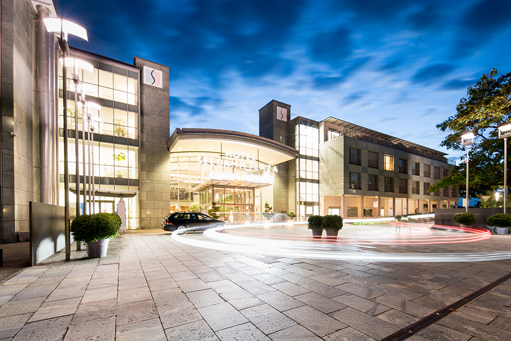 Aussenansicht des Hotels Seedamm Plaza bei Dämmerung, modern gestaltetes Gebäude mit grossem, beleuchtetem Eingangsbereich, davor ein rundes Kieselsteinpflaster mit parkendem Auto und Lichtspuren von fahrenden Fahrzeugen.