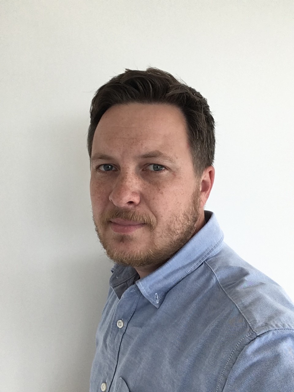 Porträt von Peter Keinz, ein Mann mit kurzem braunem Haar und Bart, der ein hellblaues Hemd trägt und vor einer weissen Wand in die Kamera blickt.