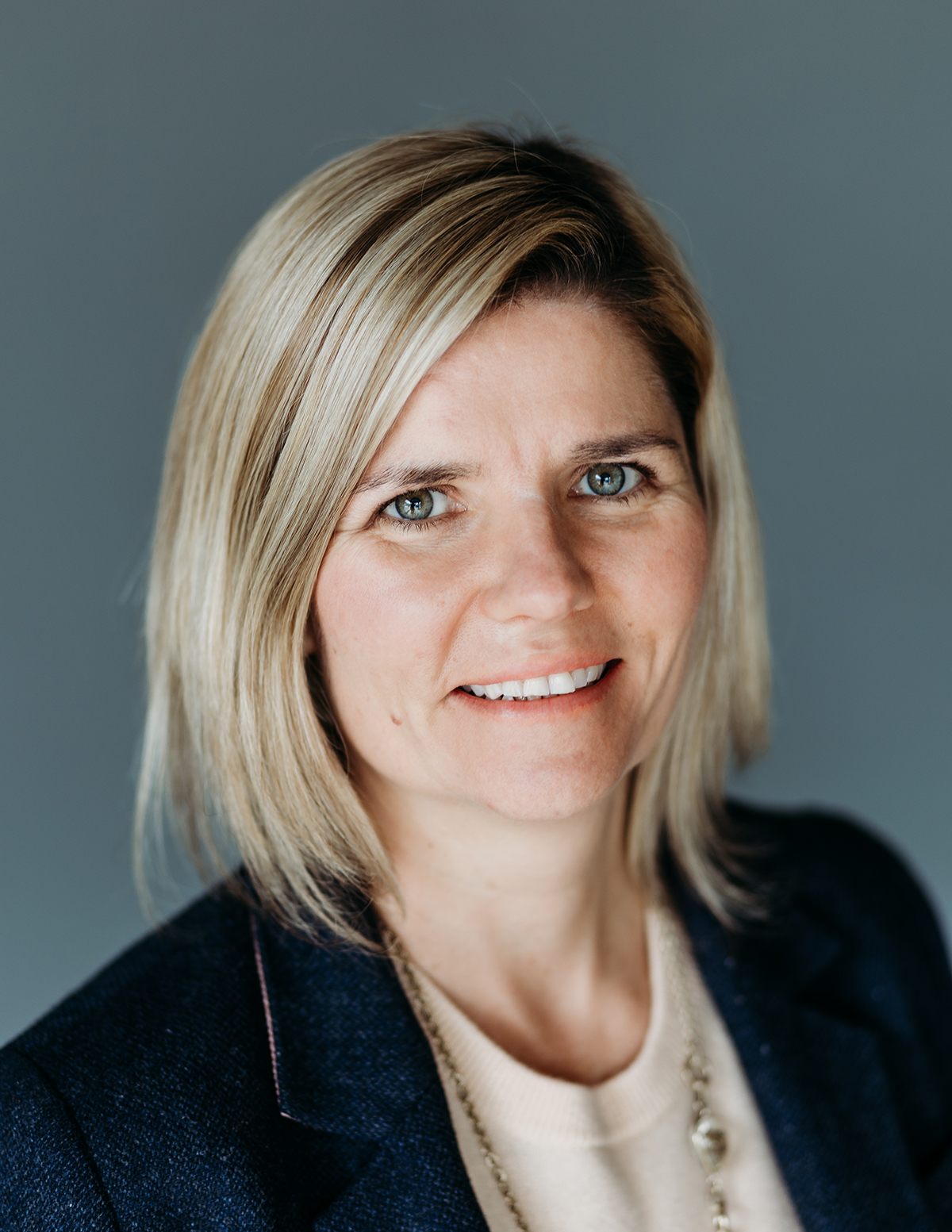 Das Porträt zeigt die Autorin mit schulterlangem blondem Haar und blauen Augen vor grauem Hintergrund. Sie trägt ein dunkelblaues Jackett über einem beigen Pullover sowie eine silberne Halskette und lächelt leicht in die Kamera.
