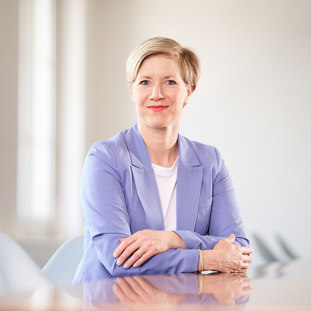 Porträt einer lächelnden Frau mit kurzem blondem Haar in einem hellvioletten Blazer und weissem Shirt, die mit verschränkten Armen an einem Konferenztisch sitzt. Der Hintergrund ist hell und unscharf mit modernen Stühlen.