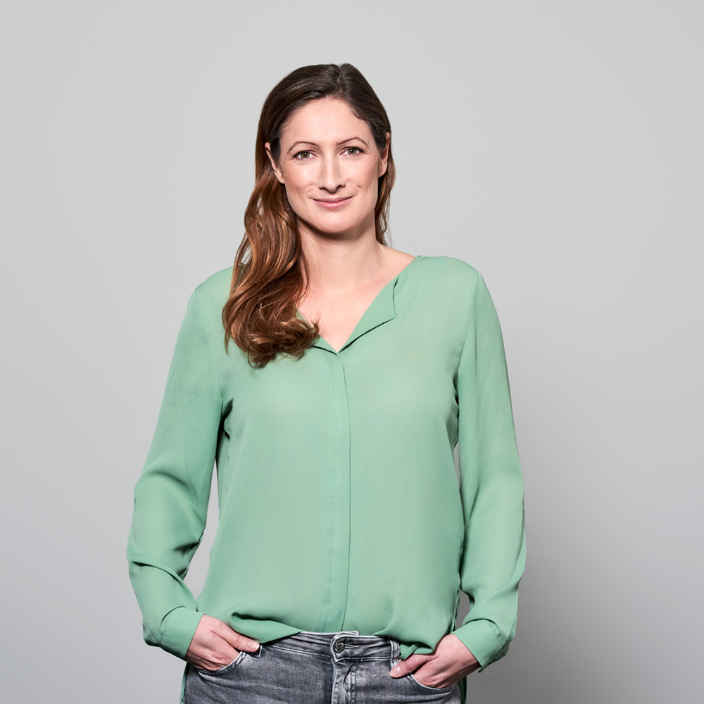 Frau mit langen, gewellten, braunen Haaren steht vor einem hellgrauen Hintergrund. Sie trägt eine mintgrüne Bluse und graue Jeans und hat die Hände locker in den Hosentaschen. Sie blickt freundlich in die Kamera.