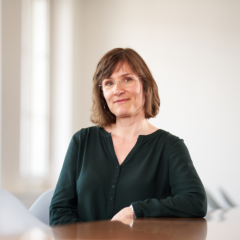Porträt einer Frau mit schulterlangem braunem Haar und Brille, die in einem dunkelgrünen Oberteil an einem Konferenztisch sitzt. Sie blickt freundlich in die Kamera, der Hintergrund ist hell und leicht verschwommen mit modernen Stühlen.