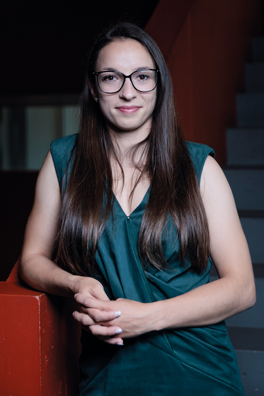 Porträt einer jungen Frau mit langen, glatten, braunen Haaren und Brille, die ein ärmelloses, dunkelgrünes Kleid trägt. Sie steht vor einer Treppe mit rotem Geländer, verschränkt die Hände locker und blickt mit leichtem Lächeln in die Kamera. Die Lichtstimmung ist gedämpft.