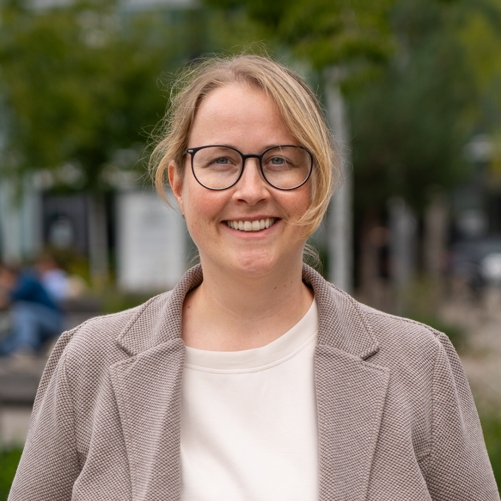 Ein Porträtfoto von Andrea Obrist im Freien. Sie ist eine Frau mit blonden, hochgesteckten Haaren und einer schwarzen Brille, die freundlich in die Kamera lächelt. Sie trägt ein helles T-Shirt unter einem strukturierten, bräunlich-grauen Blazer. Der Hintergrund zeigt einen leicht unscharfen Außenbereich mit Bäumen und Gebäuden.