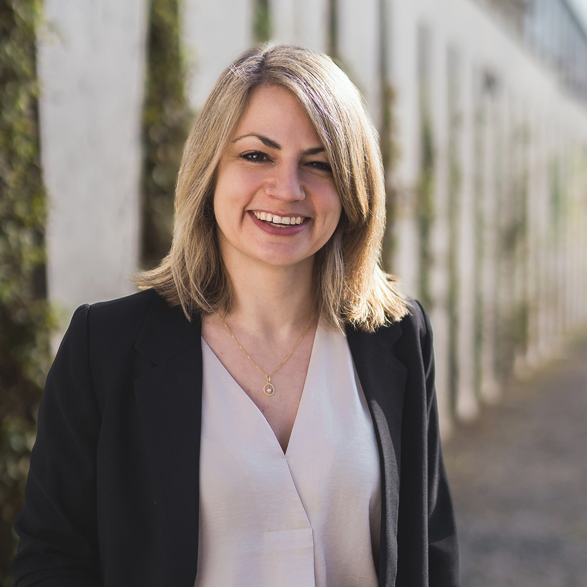 Ein Porträt von Anja Feierabend im Freien. Sie hat schulterlanges blondes Haar, lächelt freundlich in die Kamera und trägt einen schwarzen Blazer über einer hellen V-Ausschnitt-Bluse sowie eine dunkle Hose mit Gürtel. Der Hintergrund zeigt eine moderne Betonwand mit vertikalen Pflanzenelementen in selektiver Unschärfe.