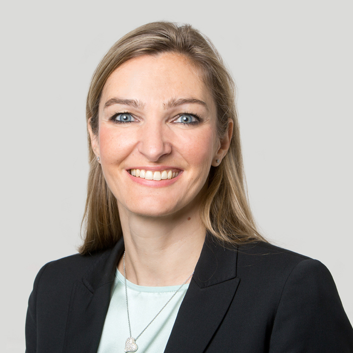 Porträt von Christine Kuhn vor einem neutralen, hellgrauen Hintergrund. Sie blickt lächelnd direkt in die Kamera. Sie hat langes, blondes Haar, blaue Augen und trägt einen schwarzen Blazer über einer helltürkisen Bluse sowie eine dezente Halskette mit einem Herzanhänger.