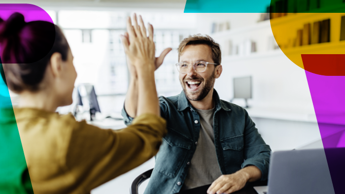 Ein Foto von zwei Personen in einem hellen, modernen Büro, die sich enthusiastisch ein High-Five geben. Links im Vordergrund ist eine Frau mit hochgesteckten dunklen Haaren und einem senfgelben Oberteil schräg von hinten zu sehen. Ihr gegenüber sitzt ein Mann an einem Schreibtisch mit einem Laptop. Er trägt eine Brille, einen Bart, ein graues T-Shirt und ein offenes, dunkelgrünes Hemd. Er blickt die Frau an und lacht dabei fröhlich.
