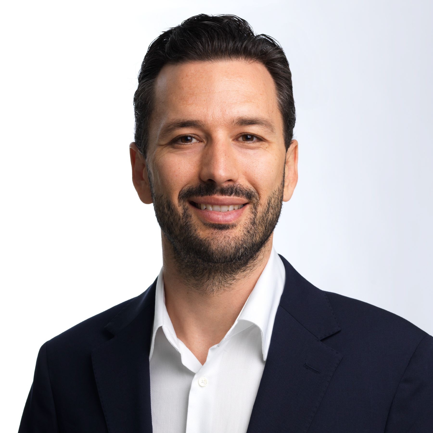 Porträt von Simon Michel. Er trägt ein dunkles Sakko über einem offenen weißen Hemd, hat dunkles, nach hinten gekämmtes Haar sowie einen gepflegten Bart und blickt lächelnd direkt in die Kamera vor einem hellen, neutralen Hintergrund.