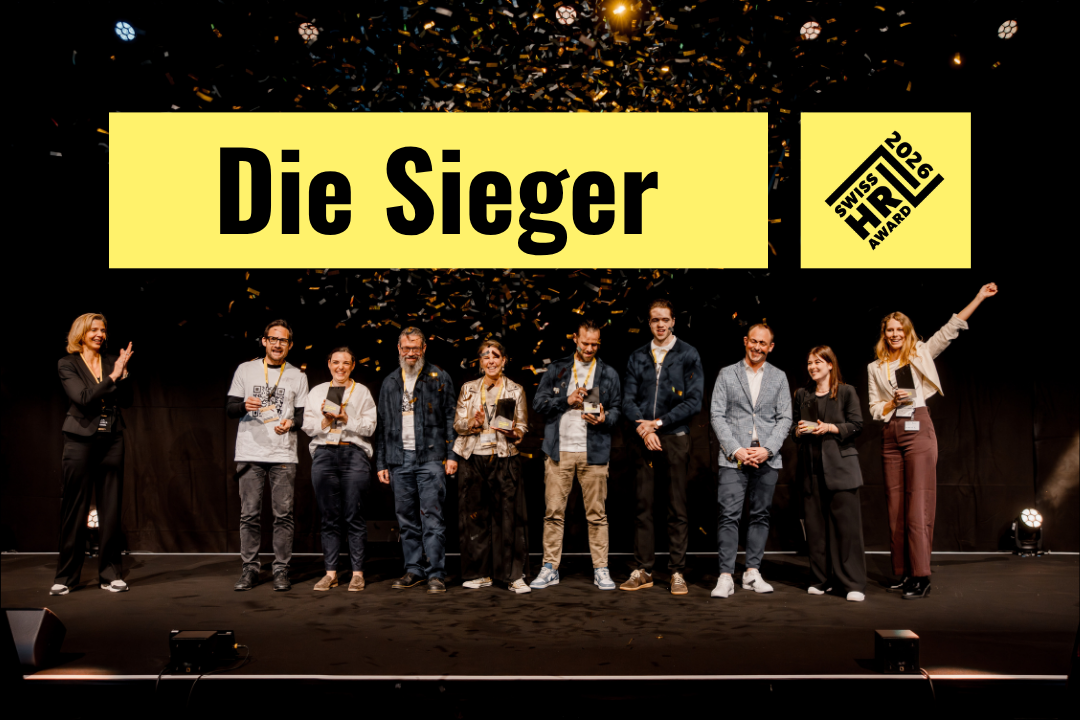 Eine Gruppe von glücklichen Gewinnern steht mit ihren schwarzen Trophäen auf einer Bühne, während goldenes Konfetti von der Decke regnet. Links im Bild steht eine Moderatorin, die den Gewinnern applaudiert.