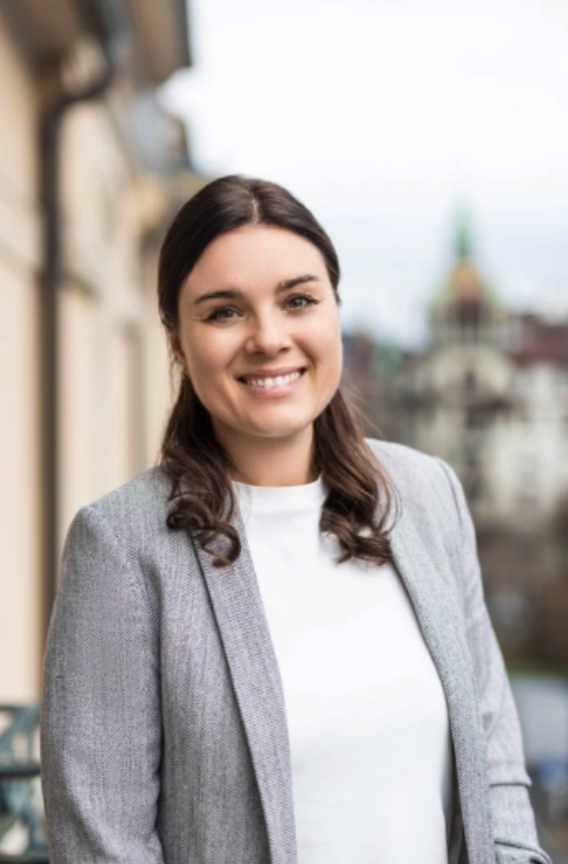 Porträt einer lächelnden Frau mit dunklem, teilweise zurückgebundenem Haar. Sie trägt einen hellgrauen Blazer über einem schlichten weißen Oberteil. Der Hintergrund ist unscharf und zeigt eine städtische Umgebung mit Gebäuden.
