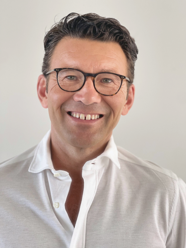 Nahaufnahme eines Mannes, der freundlich direkt in die Kamera lächelt. Er hat kurzes, dunkles und leicht gewelltes Haar, trägt eine braun gerahmte Brille und ein weißes, oben aufgeknöpftes Hemd. Der Hintergrund ist schlicht und hellgrau.