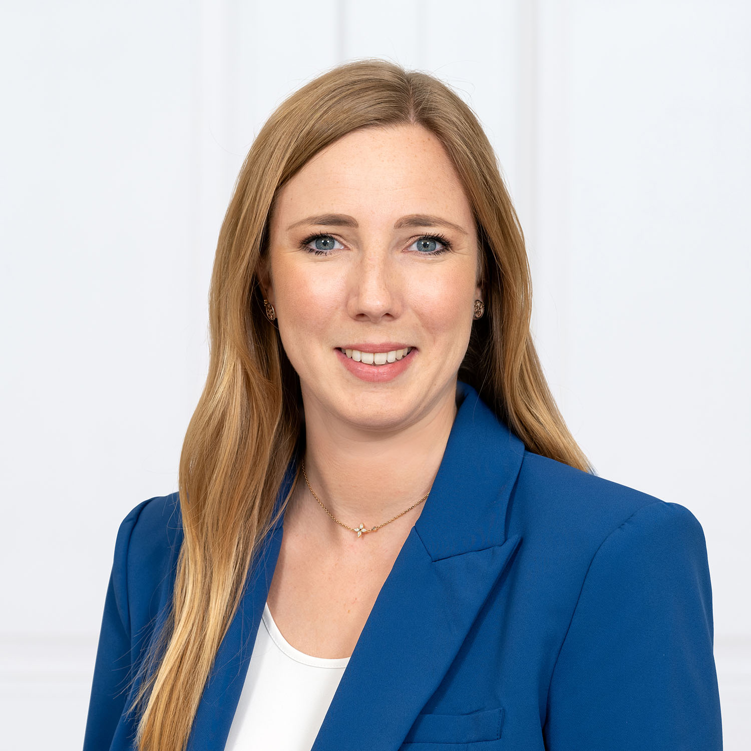 Porträtfoto einer freundlich lächelnden Frau, die direkt in die Kamera blickt. Sie hat lange, glatte blonde Haare und trägt einen leuchtend blauen Blazer über einem einfachen weißen Oberteil sowie eine zarte goldene Halskette. Der Hintergrund ist schlicht und weiß.
