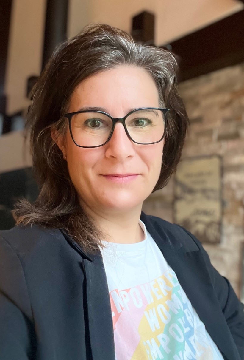 Porträt einer Frau mit schulterlangem, braun-grauem Haar und schwarzer Brille. Sie blickt mit einem leichten Lächeln direkt in die Kamera und trägt einen schwarzen Blazer über einem weißen T-Shirt mit einem bunten Schriftzug, auf dem teilweise die Worte 'EMPOWER WOMEN' zu lesen sind. Der Hintergrund ist unscharf und zeigt einen Innenraum.