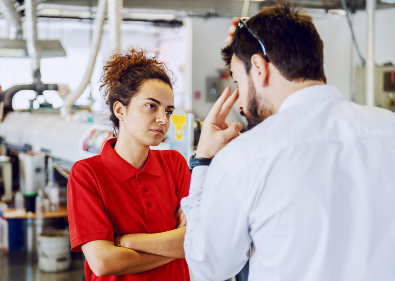 Eine junge Frau in einem roten Poloshirt steht mit verschränkten Armen in einer Werkhalle und hört mit ernstem, konzentriertem Blick einem Mann im weißen Hemd zu. Der Mann steht mit dem Rücken zur Kamera im Vordergrund und gestikuliert mit zwei Fingern auf Augenhöhe, während er auf sie einredet. Im unscharfen Hintergrund sind industrielle Maschinen und Anlagen zu erkennen.