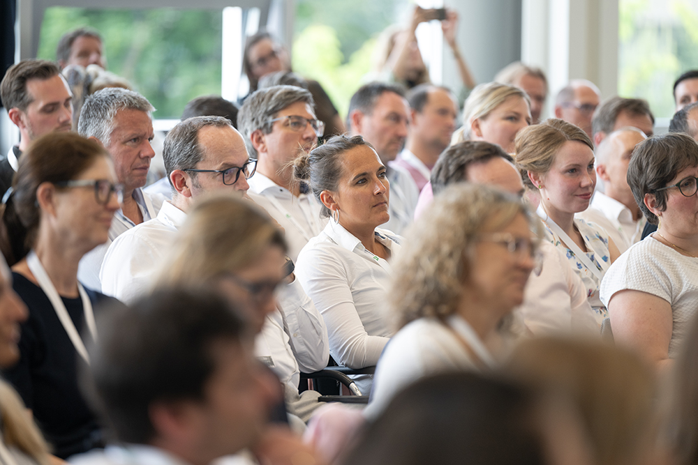 Ein dicht besetztes Publikum sitzt in den Reihen eines hellen Konferenzraums. Die Personen tragen überwiegend geschäftsmäßige Kleidung, wie helle Hemden und Blusen, und blicken aufmerksam in Richtung der Bühne. Im Hintergrund ist eine große Fensterfront mit Blick ins Grüne zu erkennen.