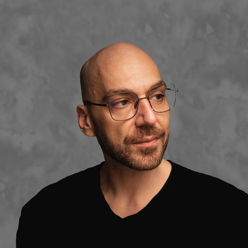 Porträt eines Mannes mit Glatze, Brille und Bart, der ein schwarzes Oberteil trägt und nach rechts aus dem Bild blickt. Der Hintergrund ist in einem strukturierten Grau gehalten.