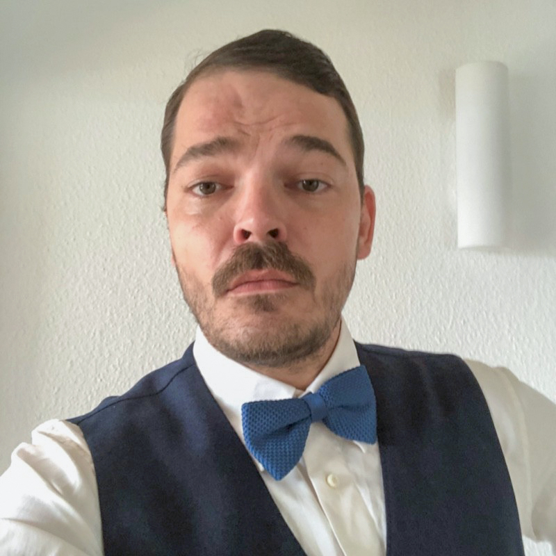 Ein Mann mit kurzem braunem Haar und Schnurrbart blickt ernst und frontal in die Kamera. Er trägt ein weißes Hemd, eine dunkelblaue Weste und eine blaue, gestrickte Fliege. Im Hintergrund ist eine weiße Wand mit einer länglichen Wandlampe zu sehen.