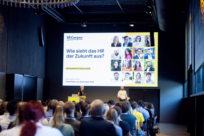 Blick von hinten über die Köpfe des Publikums auf die Bühne einer Veranstaltung. Auf einer großen Leinwand ist das Logo von „HR Campus“ sowie der Titel „Wie sieht das HR der Zukunft aus?“ und Porträts mehrerer Personen zu sehen. Vor der Leinwand stehen zwei Moderatoren auf der Bühne.