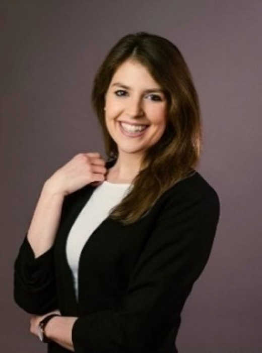 Porträt von Katharina Vögl-Duschek. Sie lächelt in die Kamera, trägt einen schwarzen Blazer über einem weissen Oberteil und hat die langen braunen Haare offen über die rechte Schulter gelegt. Hintergrund in gedecktem Graulila.