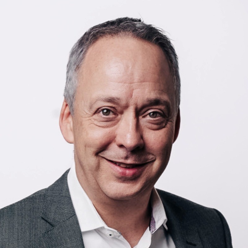 Porträt von Matthias Langenbacher. Er lächelt in die Kamera, trägt ein weisses Hemd ohne Krawatte unter einem grauen Sakko und hat kurze graumelierte Haare. Hintergrund in Weiss.