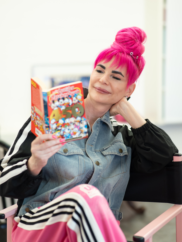 Eine Frau mit leuchtend pinkem, zu einem hohen Dutt gestylten Haar liest entspannt in einem Disney „Lustiges Taschenbuch“ (Band 450). Sie stützt ihren Kopf auf die Hand und trägt ein Outfit aus einer Jeans-Trainingsjacke und einer rosa Sporhose mit weißen Streifen.