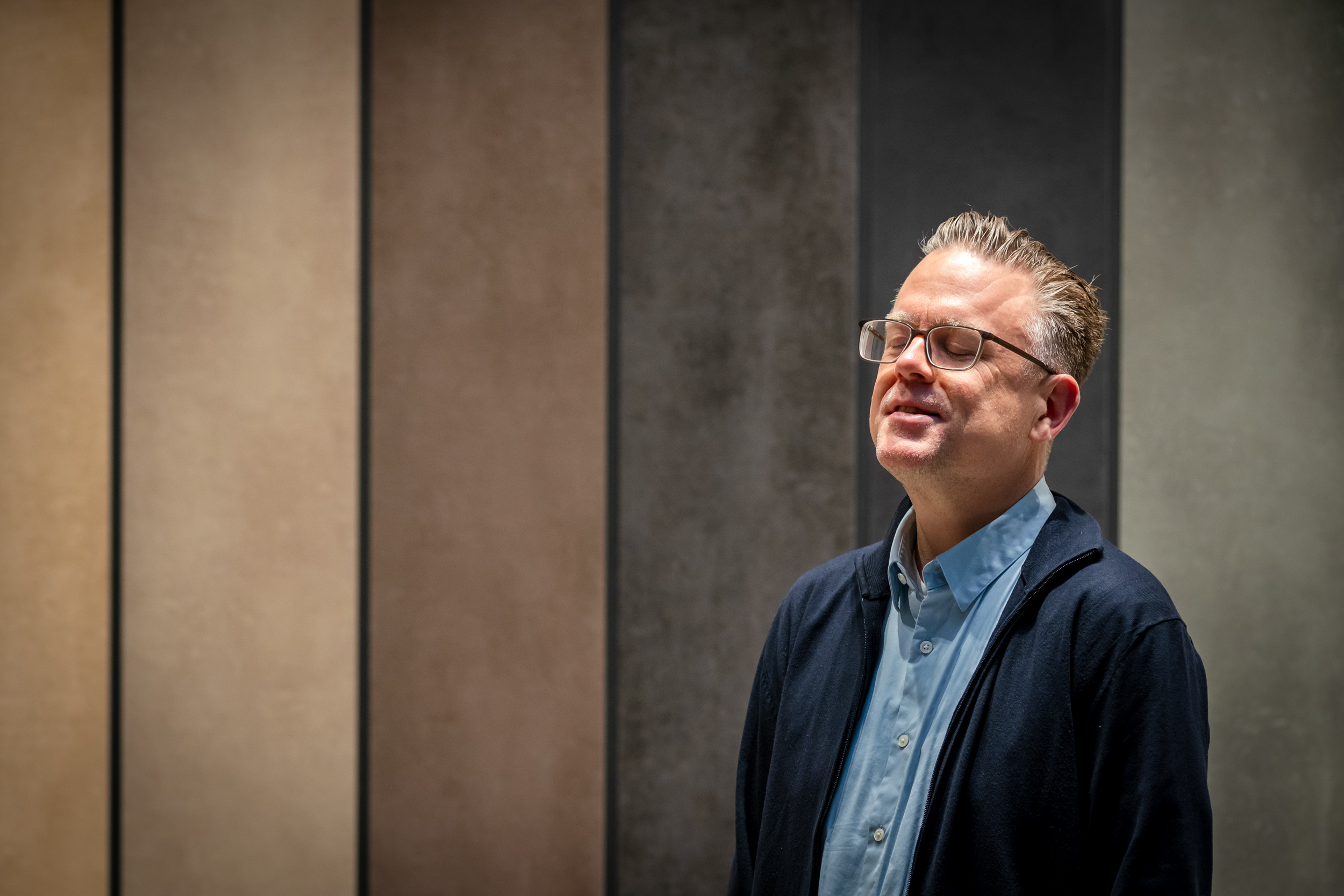 Ein Mann mit Brille, hellblauem Hemd und dunkelblauer Strickjacke steht mit geschlossenen Augen und leicht lächelndem Gesichtsausdruck vor einer Wand aus grossformatigen Materialplatten in verschiedenen Beige-, Braun- und Grautönen. Die Szene wirkt ruhig und nachdenklich.