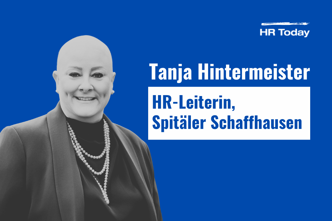 Das Bild zeigt HR-Leiterin Tanja Hintermeister in schwarz-weiss vor blauem Hintergrund. Sie hat eine Glatze, ist geschminkt und lächelt. Sie trägt einen Blazer, darunter einen Pullover und eine Perlenkette.