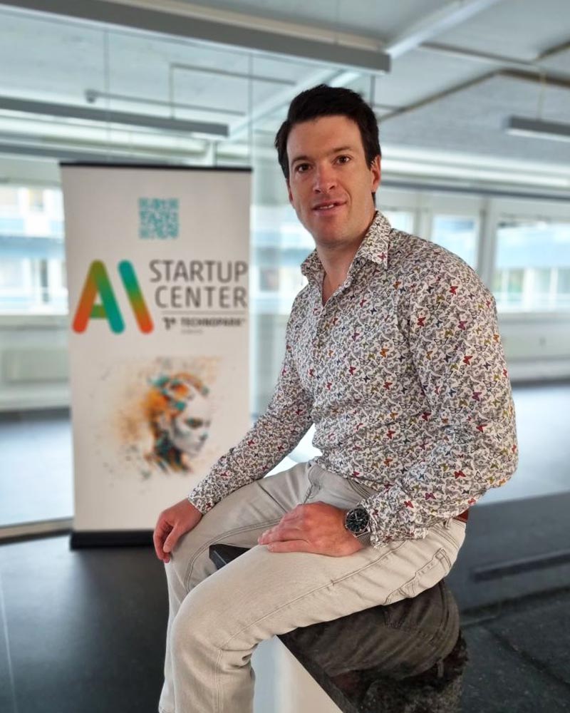 Christian Hugo Hoffmann, ein Mann mit dunklen Haaren, sitzt lächelnd im Vordergrund in einem hellen Raum mit großen Fenstern. Er trägt ein weißes Hemd mit bunten Schmetterlingsmotiven, eine helle Jeans und eine Armbanduhr. Im Hintergrund steht ein Roll-up-Banner mit dem Logo und Text "AI STARTUP CENTER", einem QR-Code und einer abstrakten, farbenfrohen Grafik eines Gesichts.