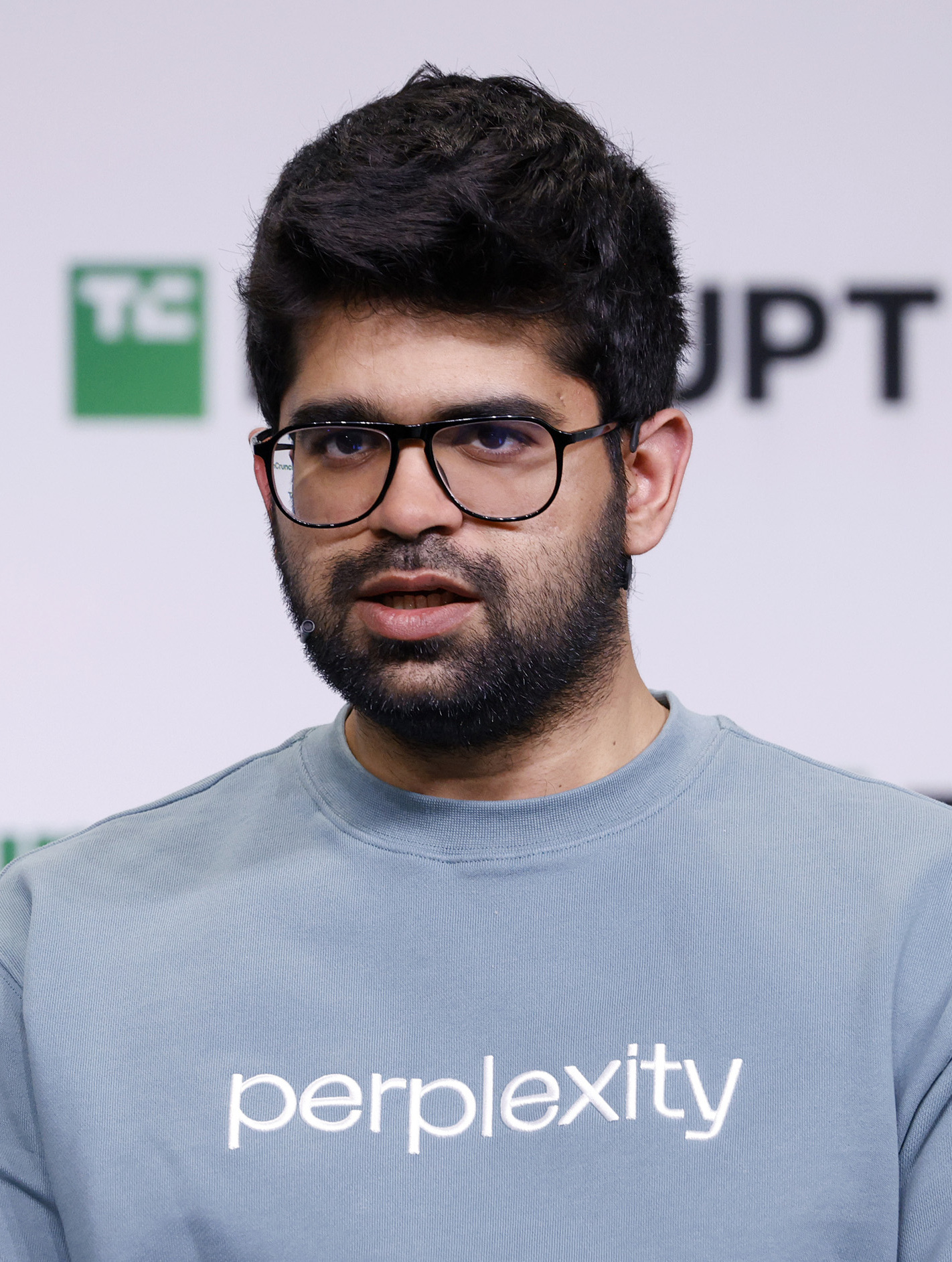 Aravind Srinivas, CEO von Perplexity AI, bei einem öffentlichen Auftritt mit blauem Sweatshirt mit „perplexity“-Schriftzug, vor einer weißen Wand mit dem TechCrunch-Logo im Hintergrund.