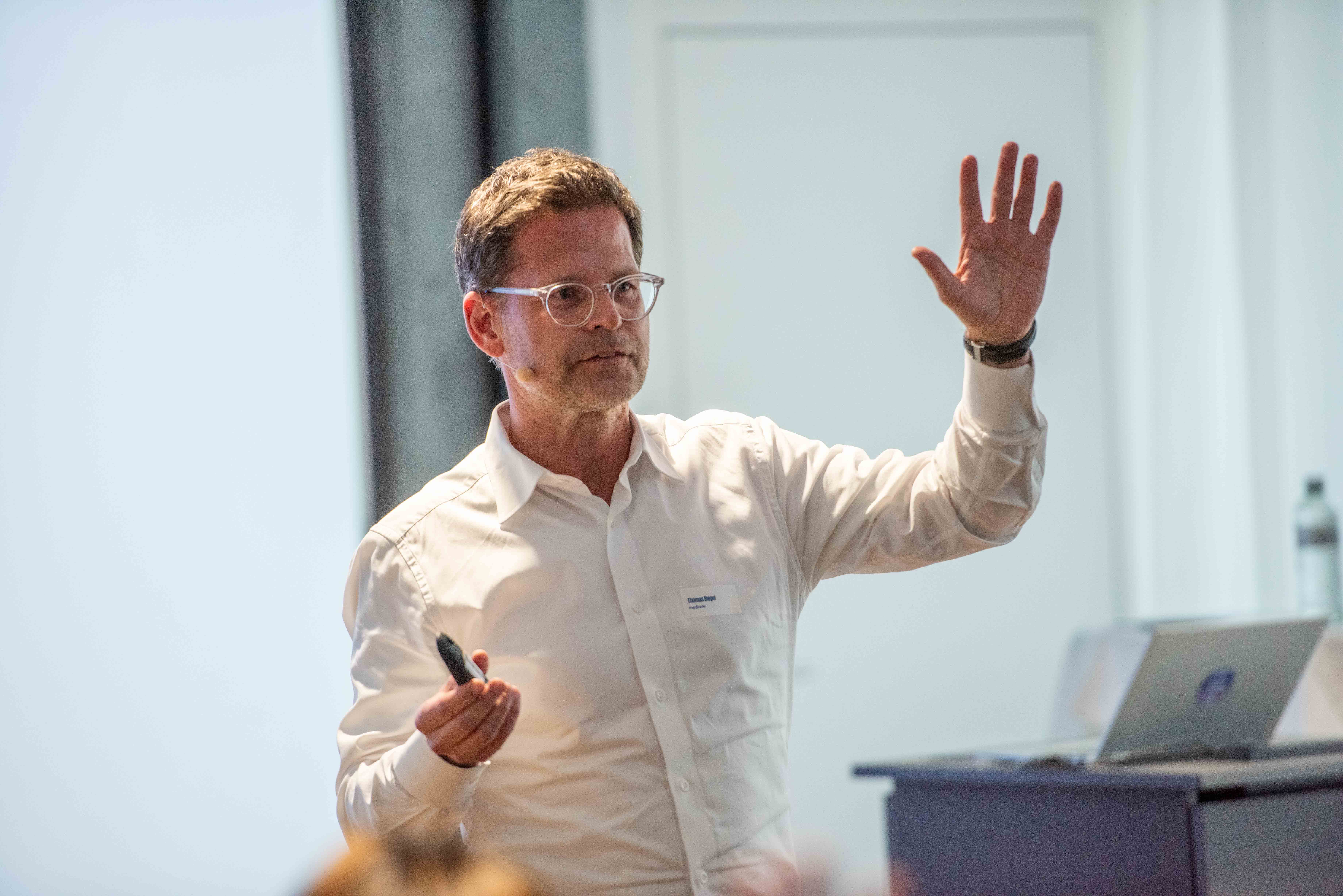 Thomas Biegel, ein Mann mit Brille in einem weißen Hemd spricht während eines Vortrags. Er hält einen Presenter in der rechten Hand und hat die linke Hand erklärend erhoben. Er trägt ein dezentes Headset-Mikrofon; im Hintergrund ist ein Laptop auf einem Pult erkennbar.