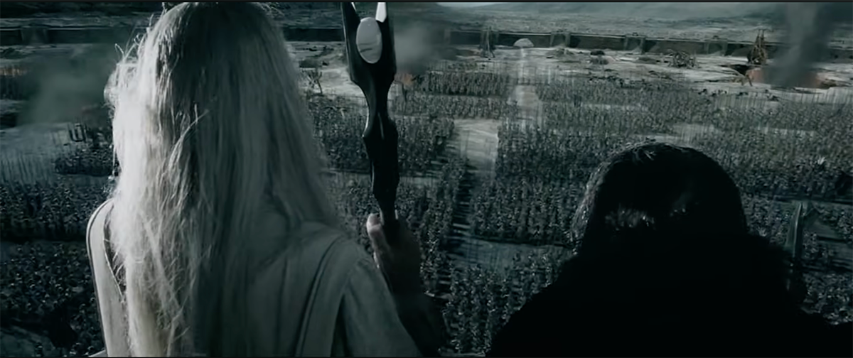 Eine Gegenüberstellung zweier Bilder mit starken visuellen Parallelen in der Darstellung von Massenaufmärschen: Das erste Bild zeigt eine Szene aus den "Herr der Ringe"-Filmen. Man sieht Saruman in weißen Roben mit seinem Stab und Gríma Schlangenzunge von hinten auf einem Balkon stehen. Sie blicken von oben auf das Industriegelände von Isengard hinab, wo eine gigantische Armee von Uruk-hai in streng geometrischen, riesigen rechteckigen Blöcken angetreten ist. Im Hintergrund steigen dunkle Rauchschwaden auf.