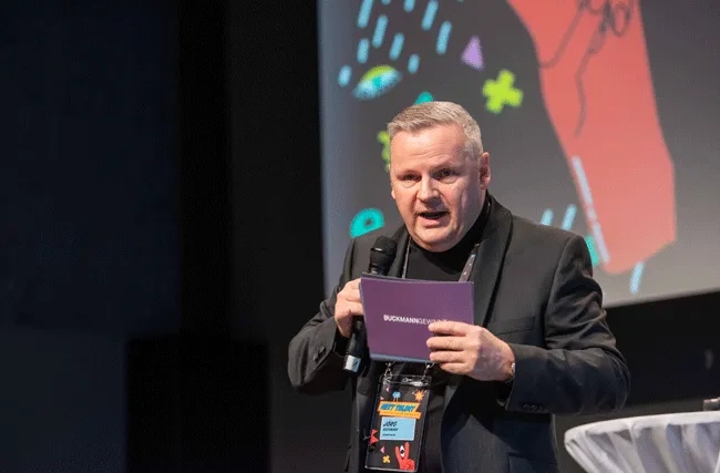 Jörg Buckmann von Buchmann Gewinnt eröffnet die Next Talent 2024. Er steht im schwarzen Anzug auf der Bühne mit Moderationskarten und Mikrofon in der Hand.
