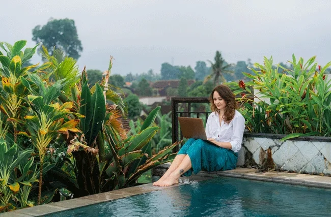 Eine Frau arbeitet am Laptop am Pool in den Tropen. Workation ermöglicht die Arbeit aus dem Ausland.