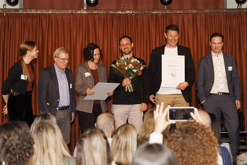Sechs Menschen von Helsana, einer mit Blumenstrauss, einer mit Gewinnerzertifikat, stehen vor Publikum auf der Bühne.