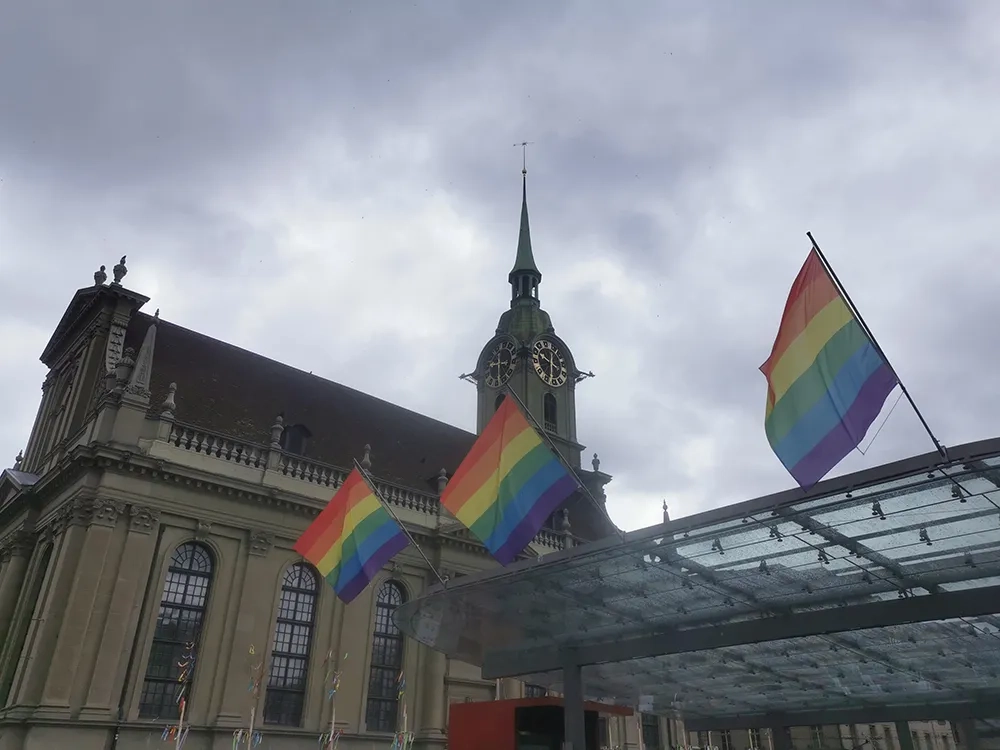 Drei Pride-Fahnen wehen im Wind am Berner Hauptbahnhof, Glockenturm im Hintergrund, bewölktes Wetter.