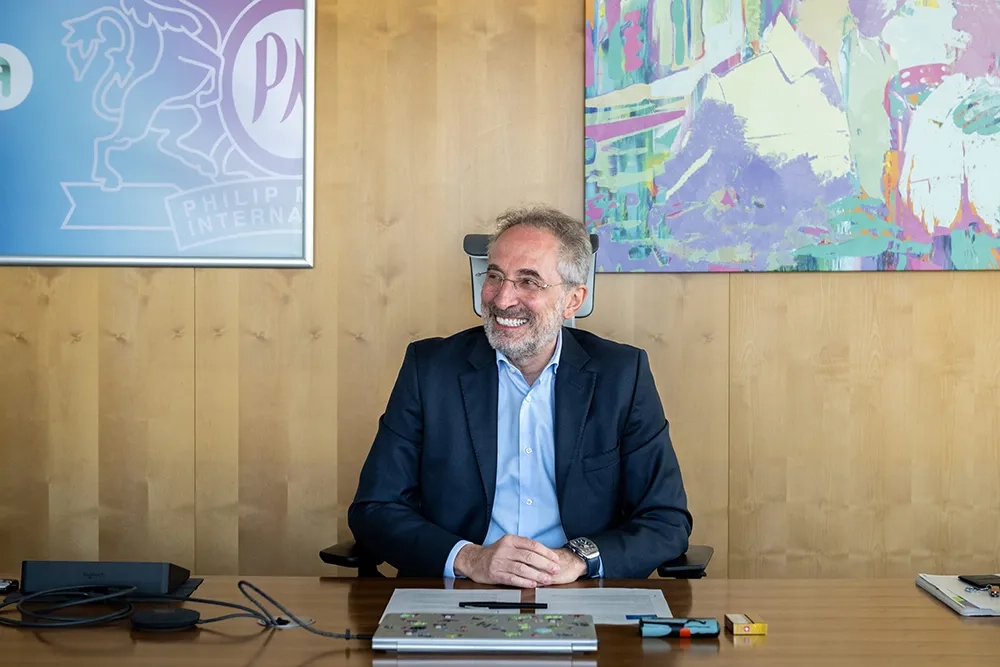 Frédéric Patitucci, Chief People & Culture Officer bei Philip Morris International, sitzt lächelnd an einem Holztisch in einem modernen Büro. Hinter ihm hängen ein farbenfrohes abstraktes Gemälde sowie ein Schild mit dem PMI-Logo. Auf dem Tisch liegen ein Laptop, ein Notizbuch und verschiedene Büromaterialien.