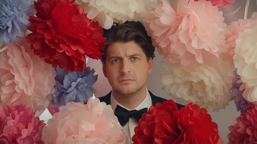 Der Schweizer Komiker Karpi blickt ernst in die Kamera, umgeben von grossen, bunten Papierblumen in Rosa, Rot, Lila und Weiß. Er trägt einen schwarzen Anzug mit Fliege. Die Szenerie wirkt übertrieben romantisch und leicht absurd – ein Kontrast zu seinem ausdruckslosen Gesichtsausdruck.