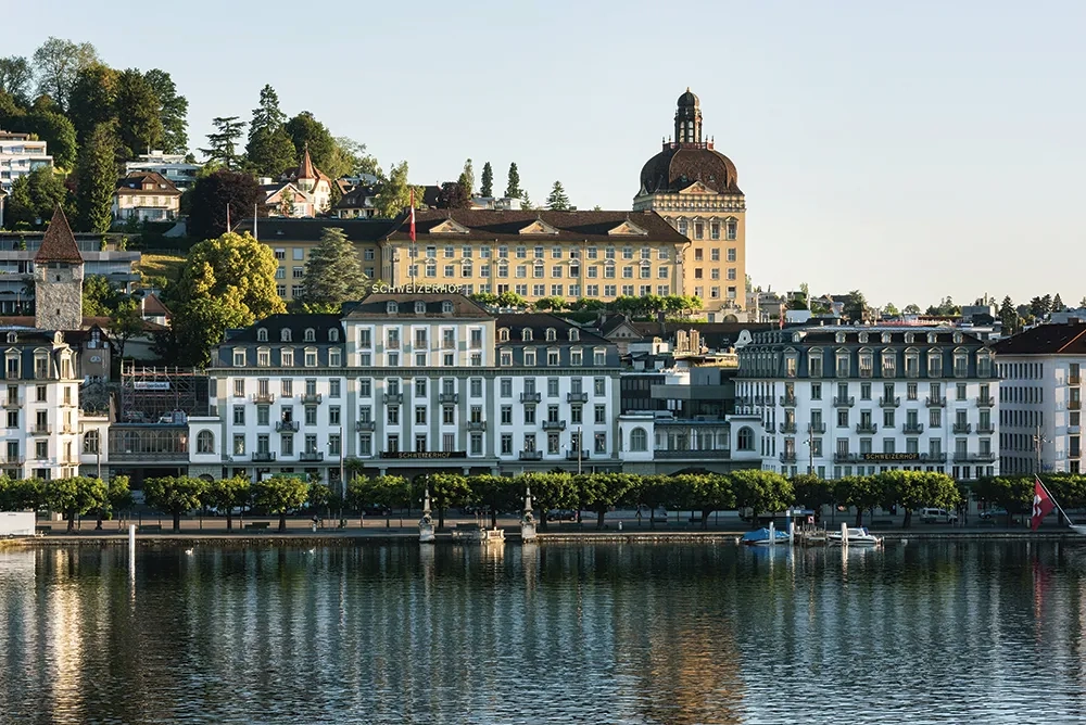 Der Hauptsitz der Suva in Luzern, aufgenommen vom gegenüberliegenden Ufer des Vierwaldstättersees. Im Vordergrund das Hotel Schweizerhof, dahinter das markante Gebäude der Suva mit seiner Kuppel und gelber Fassade, eingebettet in eine grüne Hügellandschaft.
