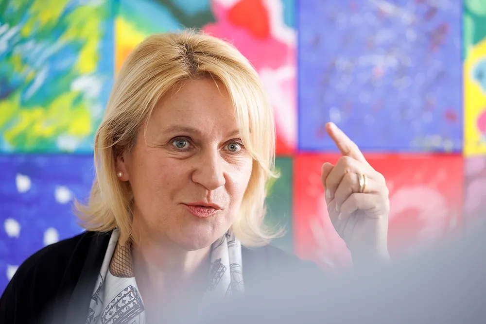 Nathalie Leschot von der Suva spricht in einem farbenfrohen Raum. Sie schaut fokussiert und hebt ihren rechten Zeigefinger, als wolle sie einen wichtigen Punkt betonen. Im Hintergrund sind bunte, abstrakte Kunstwerke zu sehen.