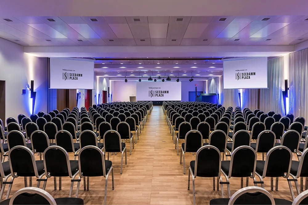 Innenaufnahme eines grossen Konferenzsaals im Seedamm Plaza, mit vielen gleichmässig angeordneten schwarzen Stühlen, einem Rednerpult auf der Bühne und drei grossen Projektionsflächen mit dem Logo des Hotels, in blauer Ambientebeleuchtung.