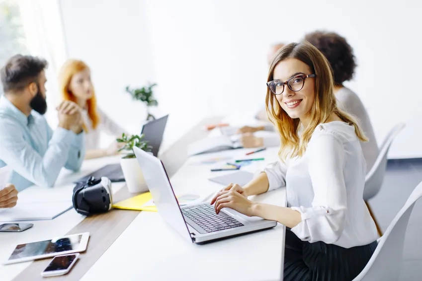 Junge Frau mit Brille sitzt an einem Konferenztisch und arbeitet lächelnd an einem Laptop. Im Hintergrund diskutieren vier weitere Personen in einem modernen, hellen Büro. Auf dem Tisch liegen elektronische Geräte, Unterlagen und Büromaterialien. 