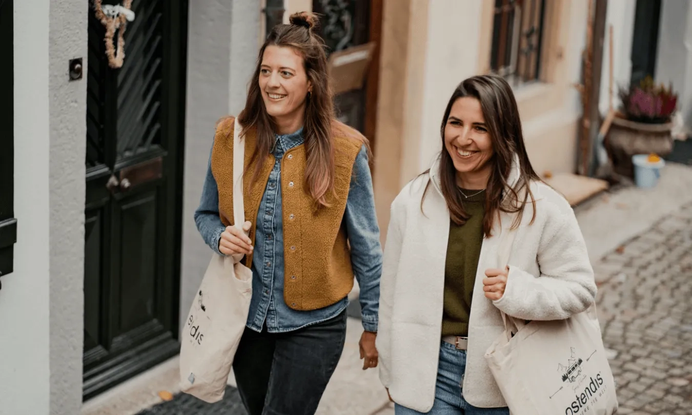 Zwei lachende Frauen spazieren nebeneinander. Die Frau auf der linken Seite hat lange braune Haare, die teilweise zu einem Dutt hochgesteckt sind. Sie trägt ein blaues Jeanshemd unter einer braun-senffarbenen Teddyfell-Weste und eine dunkle Hose. Die Frau rechts hat ebenfalls lange, dunkle Haare und trägt einen olivgrünen Pullover, eine cremefarbene Teddyfell-Jacke sowie eine blaue Jeans. Beide Frauen tragen helle Stoffbeutel mit der Aufschrift und dem Logo "ostendis".