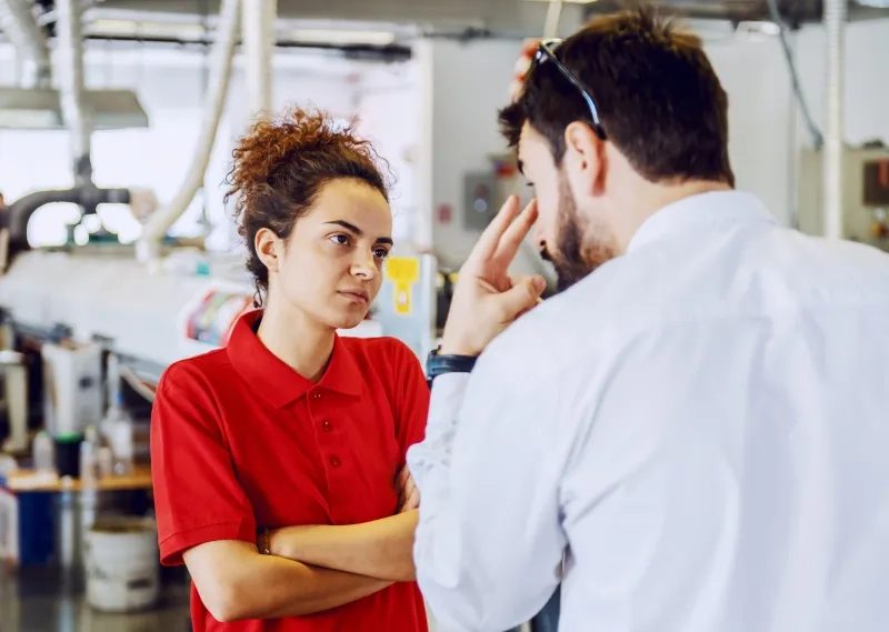 Eine junge Frau in einem roten Poloshirt steht mit verschränkten Armen in einer Werkhalle und hört mit ernstem, konzentriertem Blick einem Mann im weißen Hemd zu. Der Mann steht mit dem Rücken zur Kamera im Vordergrund und gestikuliert mit zwei Fingern auf Augenhöhe, während er auf sie einredet. Im unscharfen Hintergrund sind industrielle Maschinen und Anlagen zu erkennen.