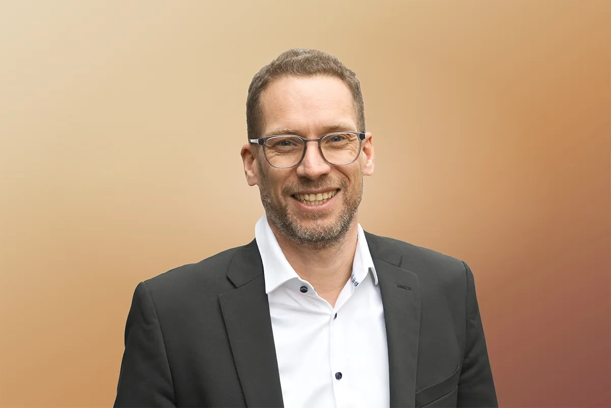 Lars Joeckel, kurze graumelierte Haare, Bart und graue Brille, lächelt in die Kamera. Er trägt ein weisses Hemd und ein dunkles Sakko, im Hintergrund ein warmer orange-beiger Farbverlauf.