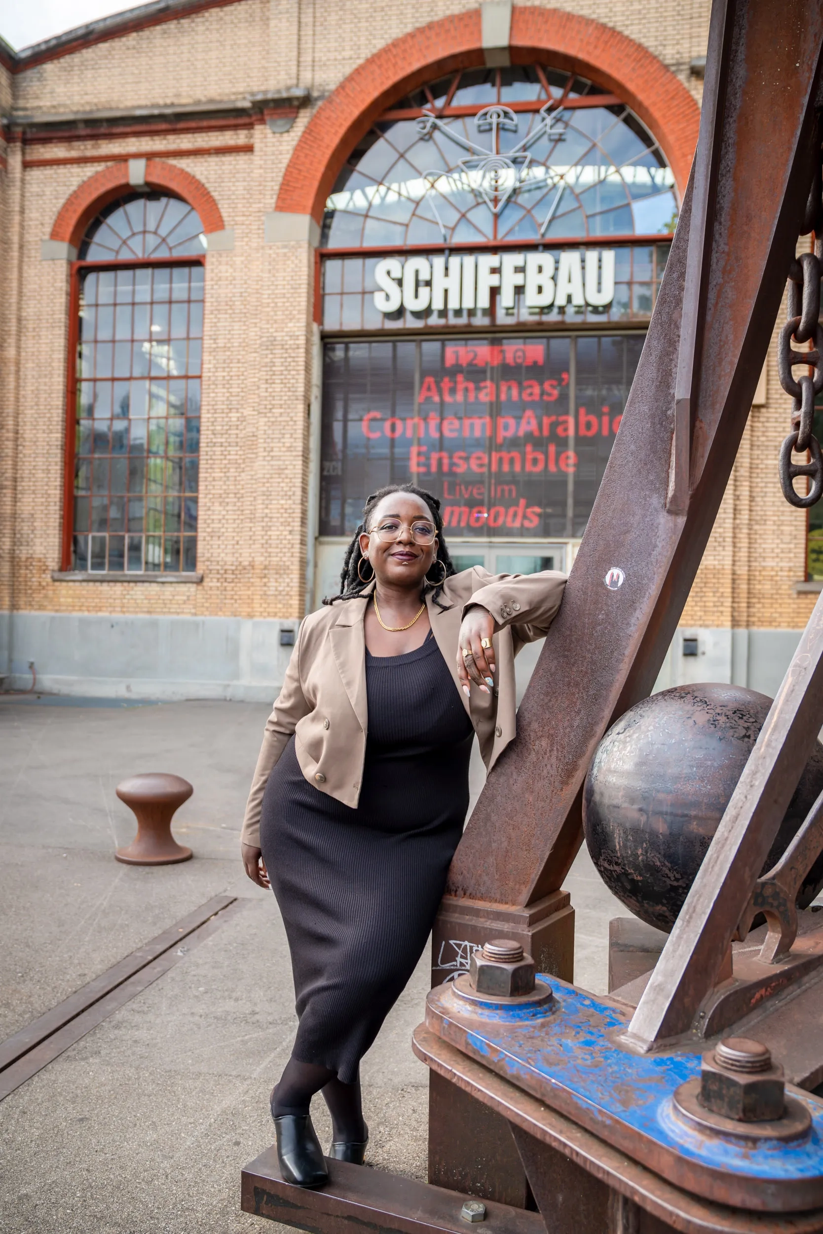 Yuvviki Dioh lehnt sich an eine Eisenskulptur, hinter ihr der Eingang zum Schiffbau. Sie trägt ein schwarzes Kleid und einen hellbraunen Blazer.
