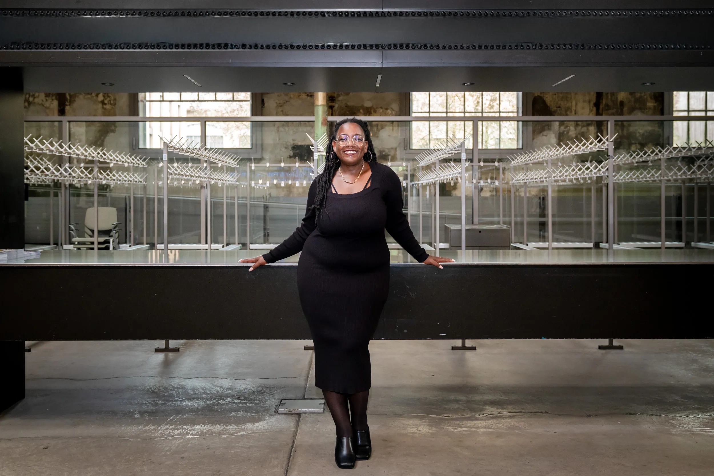 Yuvviki Dioh steht vor der leeren Garderobe im Schiffbau und lehnt an leicht an den Tresen an, ihre Arme nach aussen ausgebreitet,. Sie trägt ein schwarzes Kleid, Strümpfe und schwarze Schuhe, eine Brille und lächelt breit.